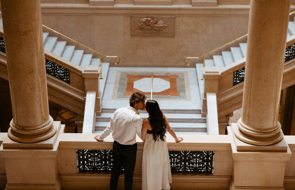 couple kissing in front of grand staircase during art museum engagement session photographed by pittsburgh engagement photographer