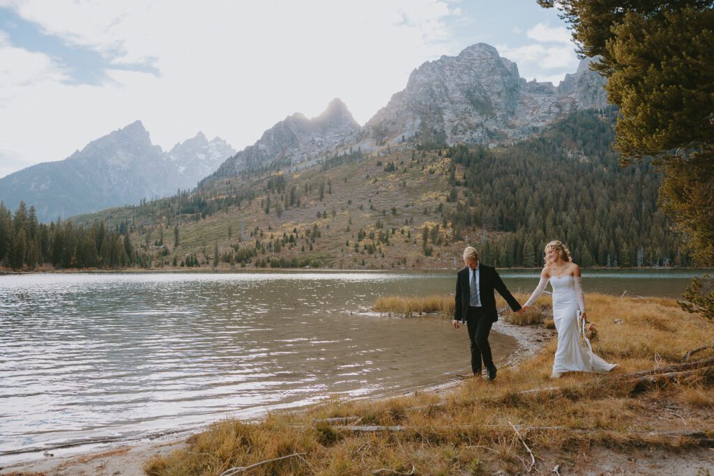 Eloping couple at sunset in Grand Teton National Park walking and holding hands in front of a lake.